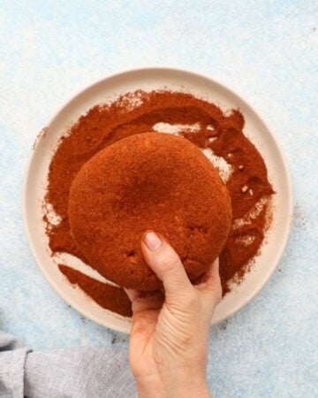 two hands rolling a pink cheese ball in paprika. 