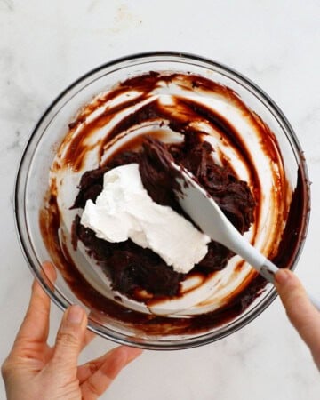 two hands mixing brown chocolate mousse in a glass bowl.
