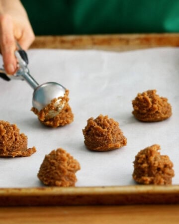 a hand adding scoops of brown ginger molasses cookie dough on a white baking sheet.
