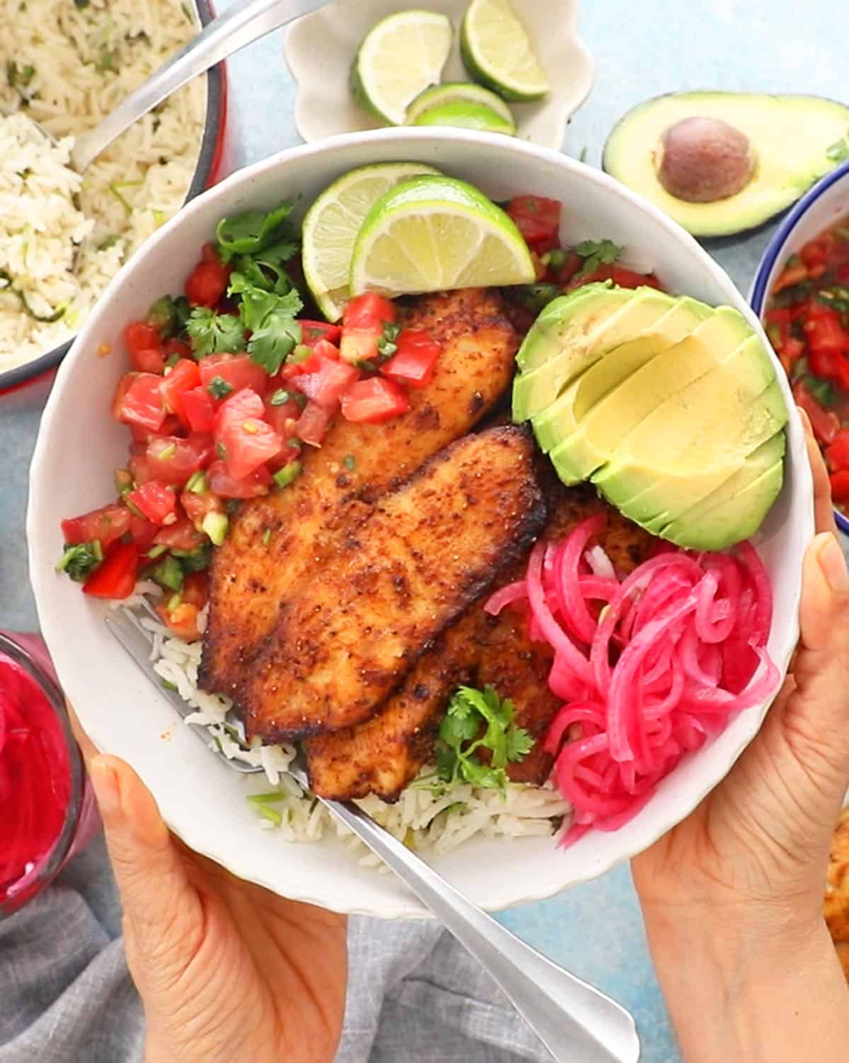 two hands holding a white bowl with brown tilapia, green avocado and pink onions.