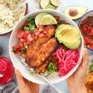 two hands holding a white bowl with brown tilapia, green avocado and pink onions.
