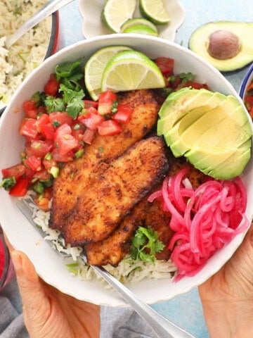 two hands holding a white bowl with brown tilapia, green avocado and pink onions.